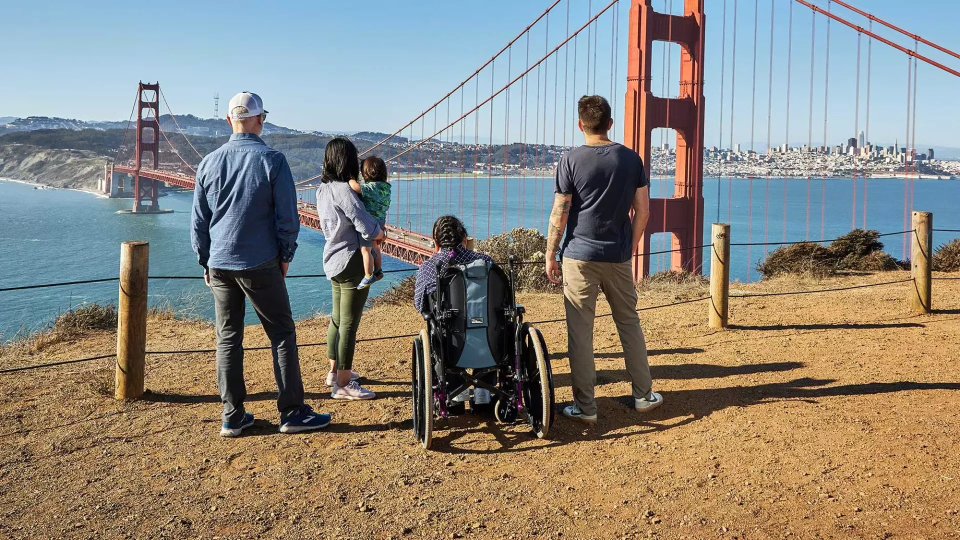 一群人, 包括一个坐轮椅的人, ist von hinten zu sehen, während sie von den Marin Headlands aus auf die Golden Gate Bridge blicken.