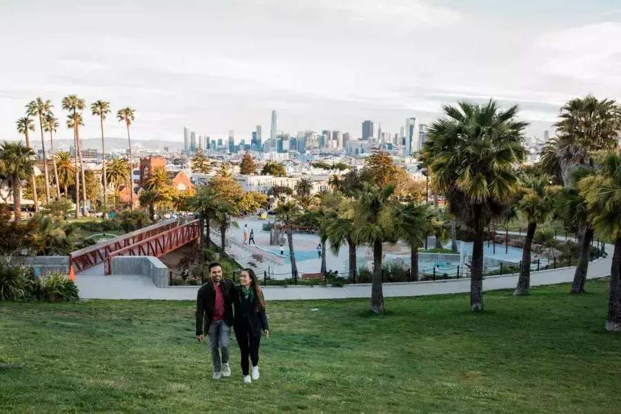 一对夫妇走向镜头，Dolores Park和贝博体彩app的天际线在他们身后.