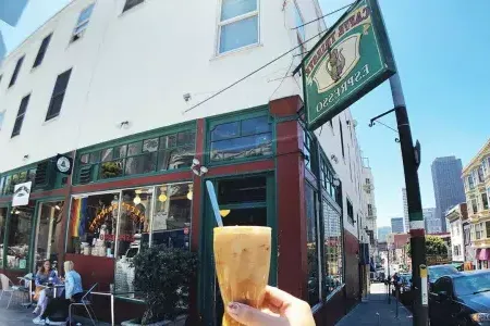 A person's hand holds a coffee drink with the exterior sign and storefront of Caffe Trieste in the background.