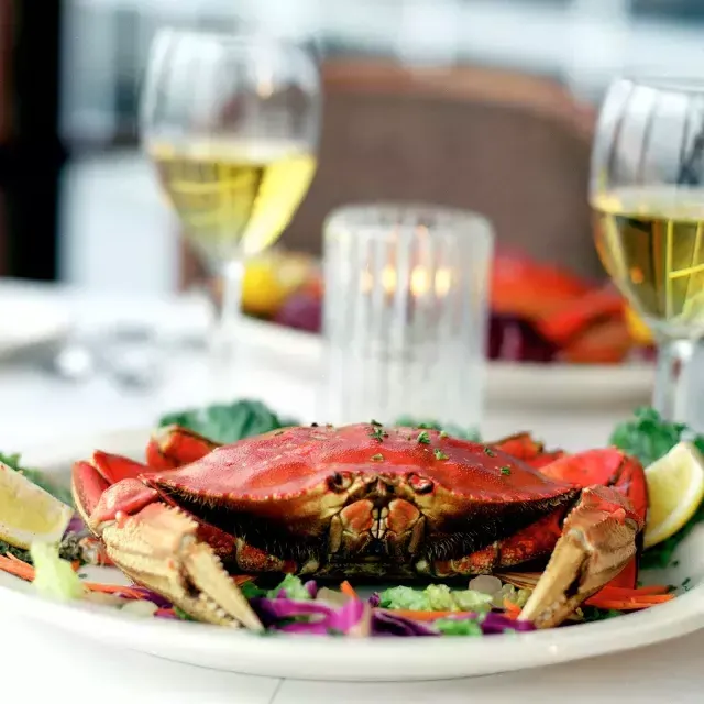 Um caranguejo Dungeness sentado em um prato em 一个餐厅 com duas taças de vinho branco ao fundo.